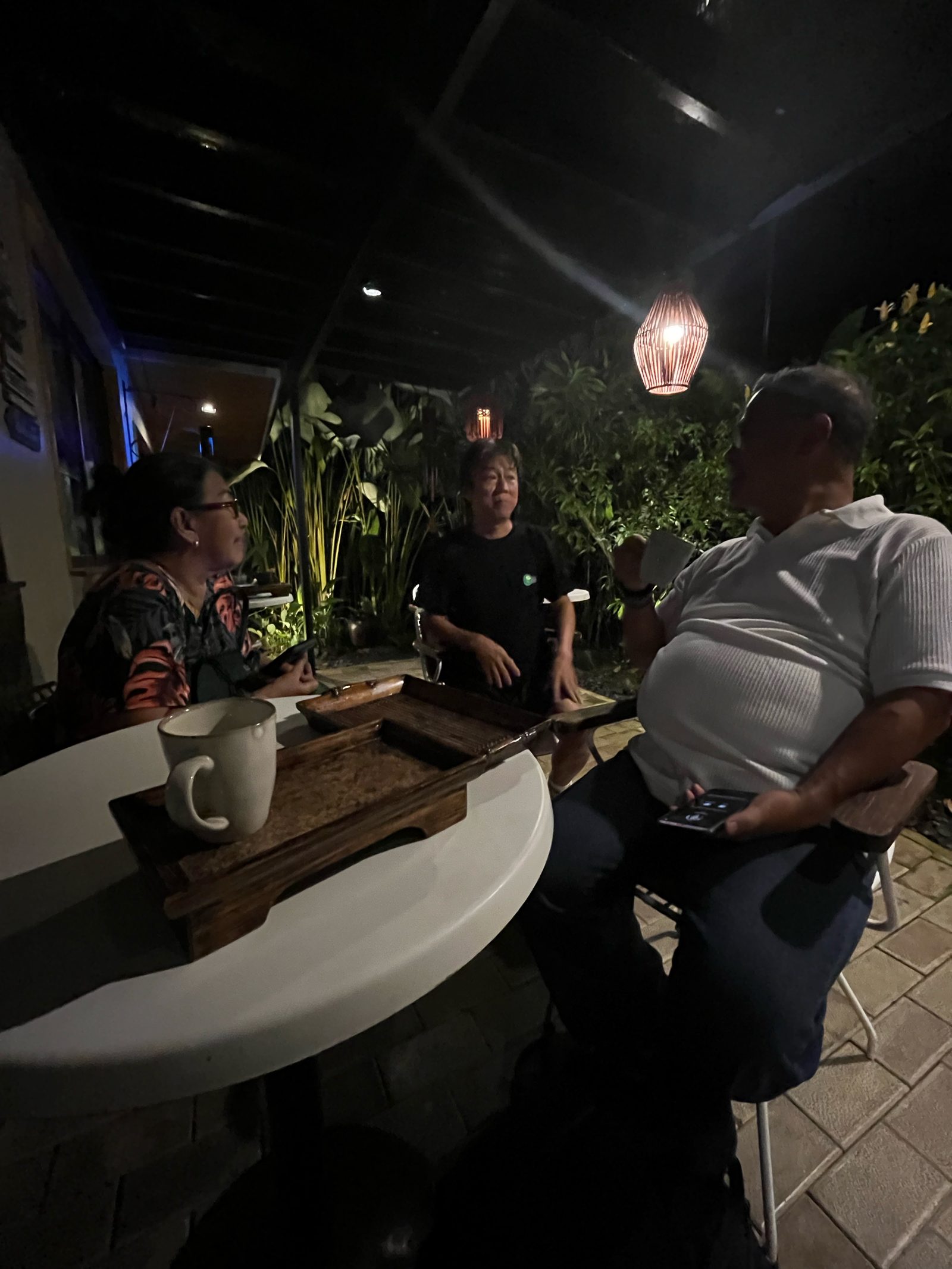 Guests at a wooden table in the Tea Garden at night, with ginger tea and woven lanterns overhead.